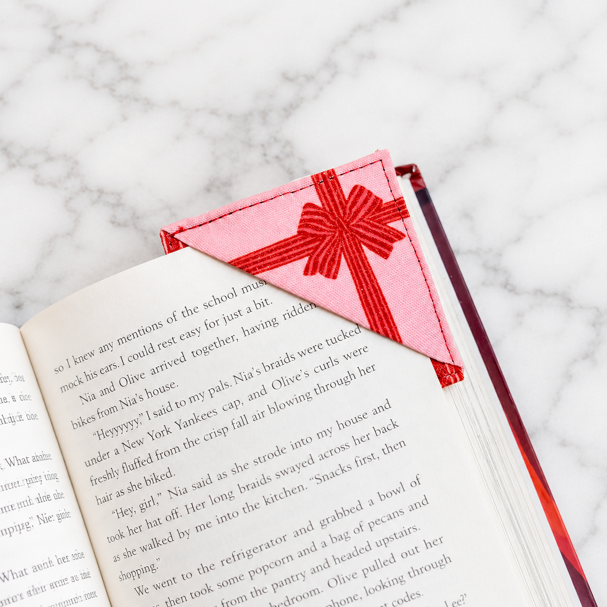 A pink fabric bookmark with a red ribbon bow and red spine, placed on an open book on a white marble surface.
