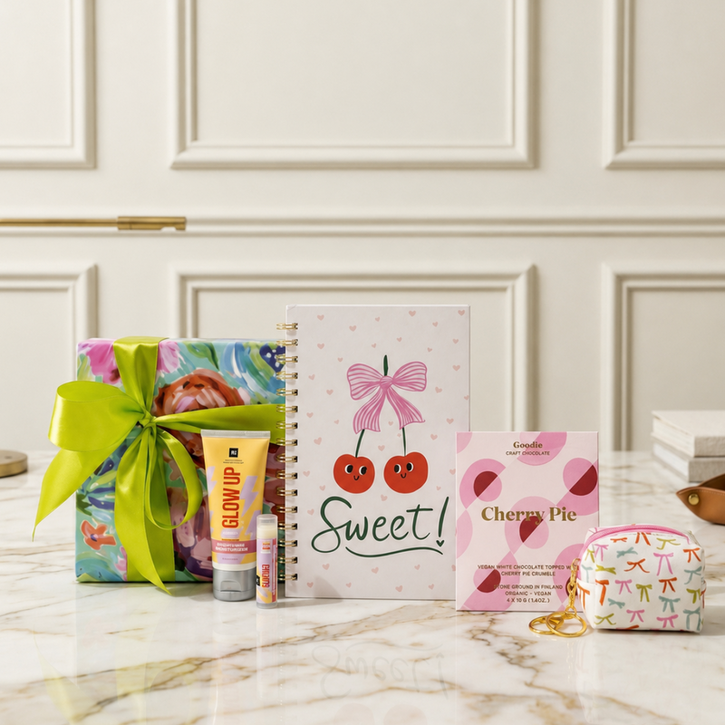 A curated stationery gift set includes a wrapped gift box with a satin ribbon, a heart designed spiral notebook with cherries and the word 'Sweet!', a hand cream, lip butter balm, a salted caramel chocolate bar and and a small bow designed keychain pouch. 

The perfect curated gift for celebrations, milestones, graduations, teens, tweens, pre-teens, stationery lovers.