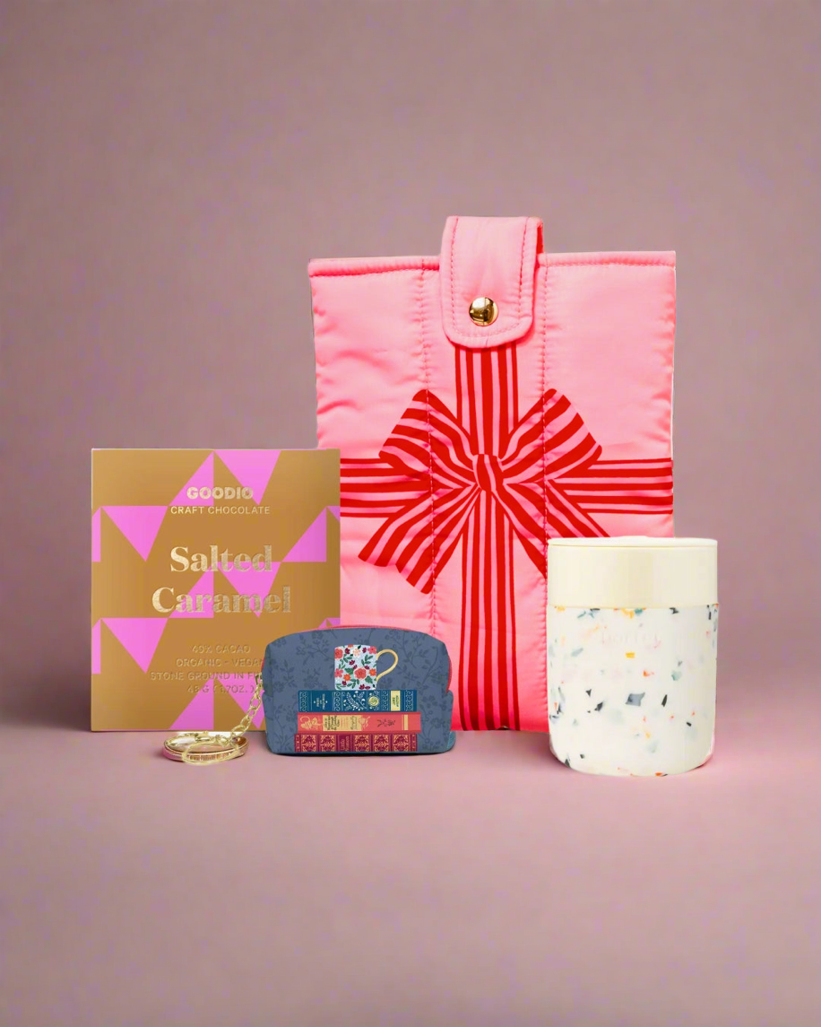 gift set which includes of pink book pouch with red bow pattern, salted caramel chocolate bar, small blue keychain pouch, and white ceramic travel mug candle on a pink background