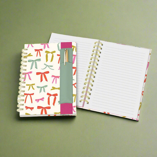 Notebook with colorful bow pattern on cover, pen insert and open to lined pages on a green background