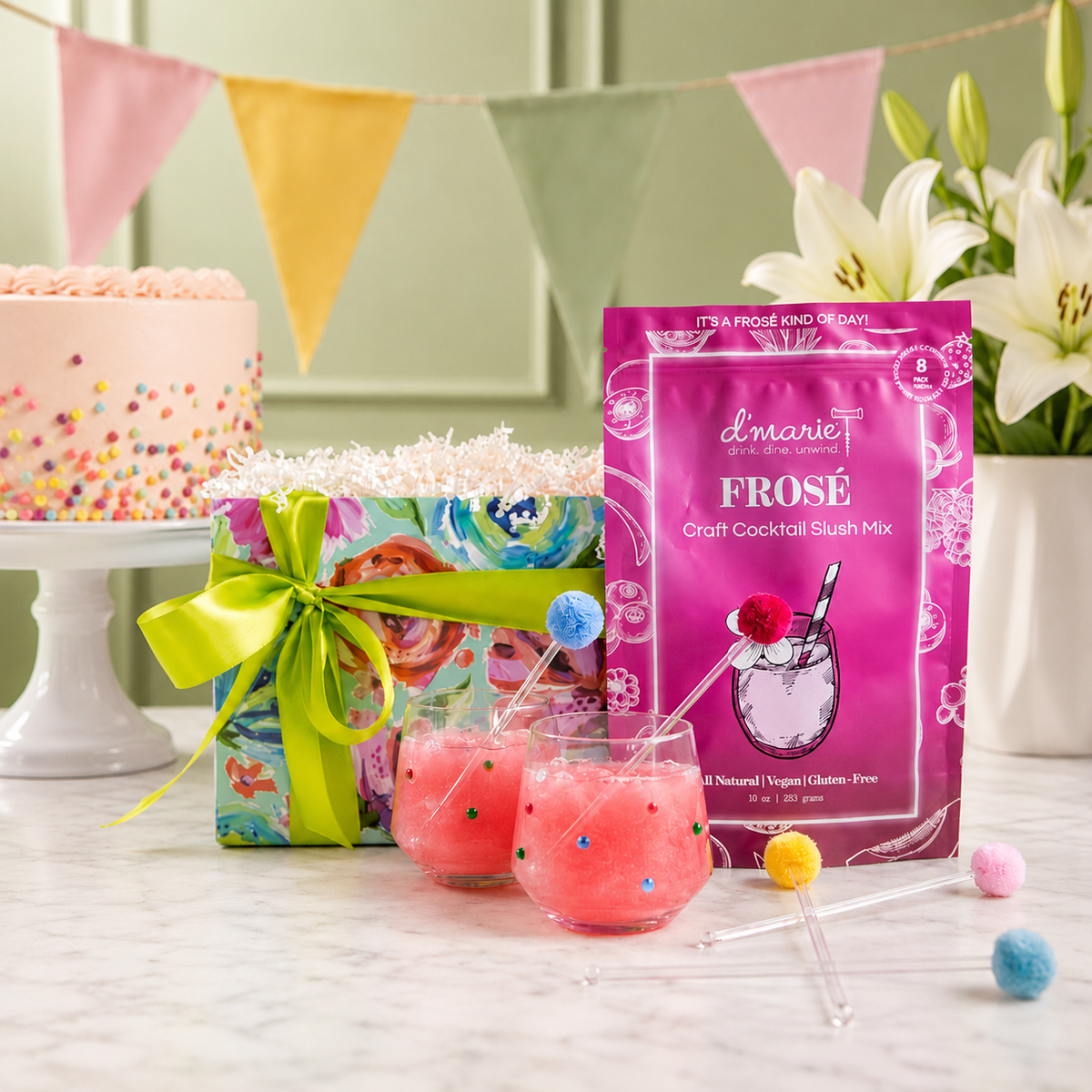 A celebration curated gift includes Frose Craft Cocktail Slush Mix, two frose slush drinks in clear confetti beaded wine glasses, and colorful pom pom drink stirrers on a marble surface. A decorated cake on a stand and white lilies are blurred in the background under a pastel bunting banner.