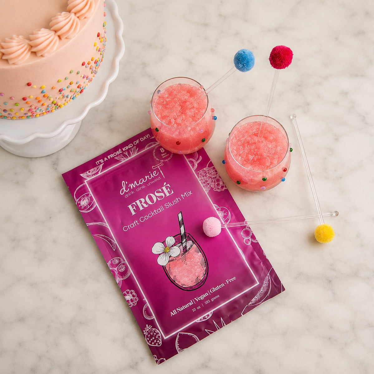 A celebration curated gift includes Frose Craft Cocktail Slush Mix, two frose slush drinks in clear confetti beaded wine glasses, and colorful pom pom drink stirrers. A decorated cake on a marble surface.