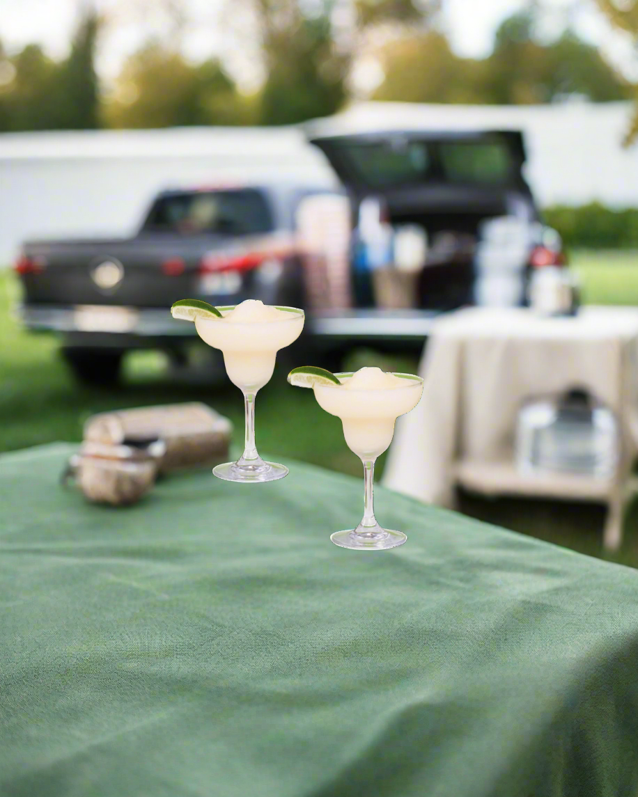 Two beergarita glasses on a green tablecloth with a blurred background of a car and people.