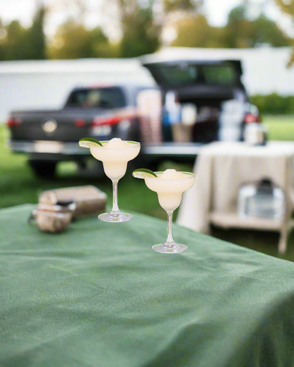 Two beergarita glasses on a green tablecloth with a blurred background of a car and people.