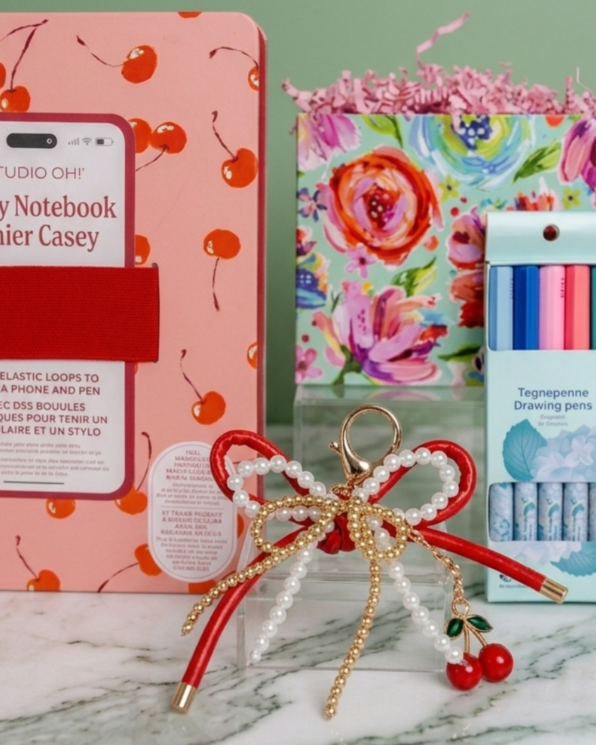 Stationery gift set including a red cherry decorative notebook, decorative gift basket box, and cherry charm bag keychain on a marble surface with a green background.