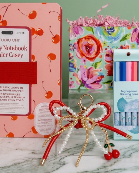 Stationery gift set including a red cherry decorative notebook, decorative gift basket box, and cherry charm bag keychain on a marble surface with a green background.