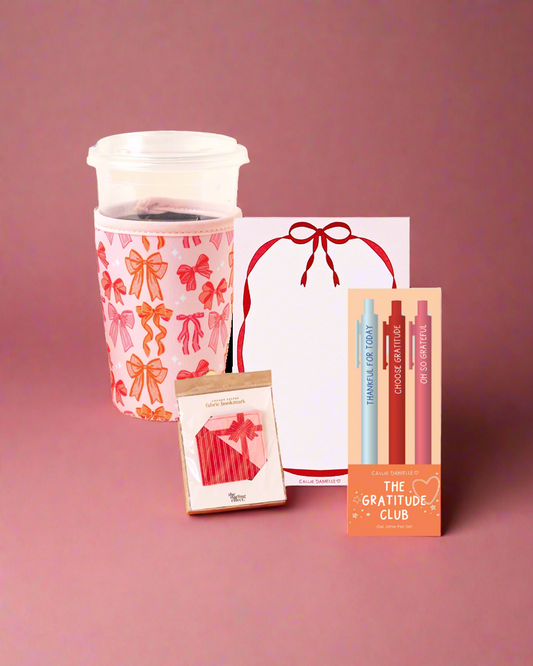 gift set including a drink sleeve with a bow pattern, a bow design notepad, a fabric bookmark, and set of pen with gratitude wording on a pink background