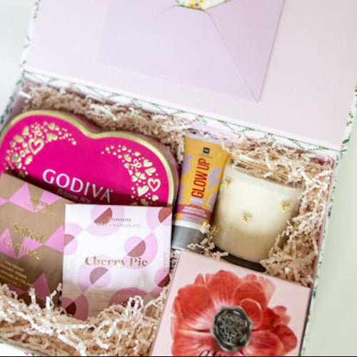An open gift box filled with assorted gift items, including a pink Godiva heart-shaped chocolate, a small candle, a hand cream, and a floral card, set against a clean white background.