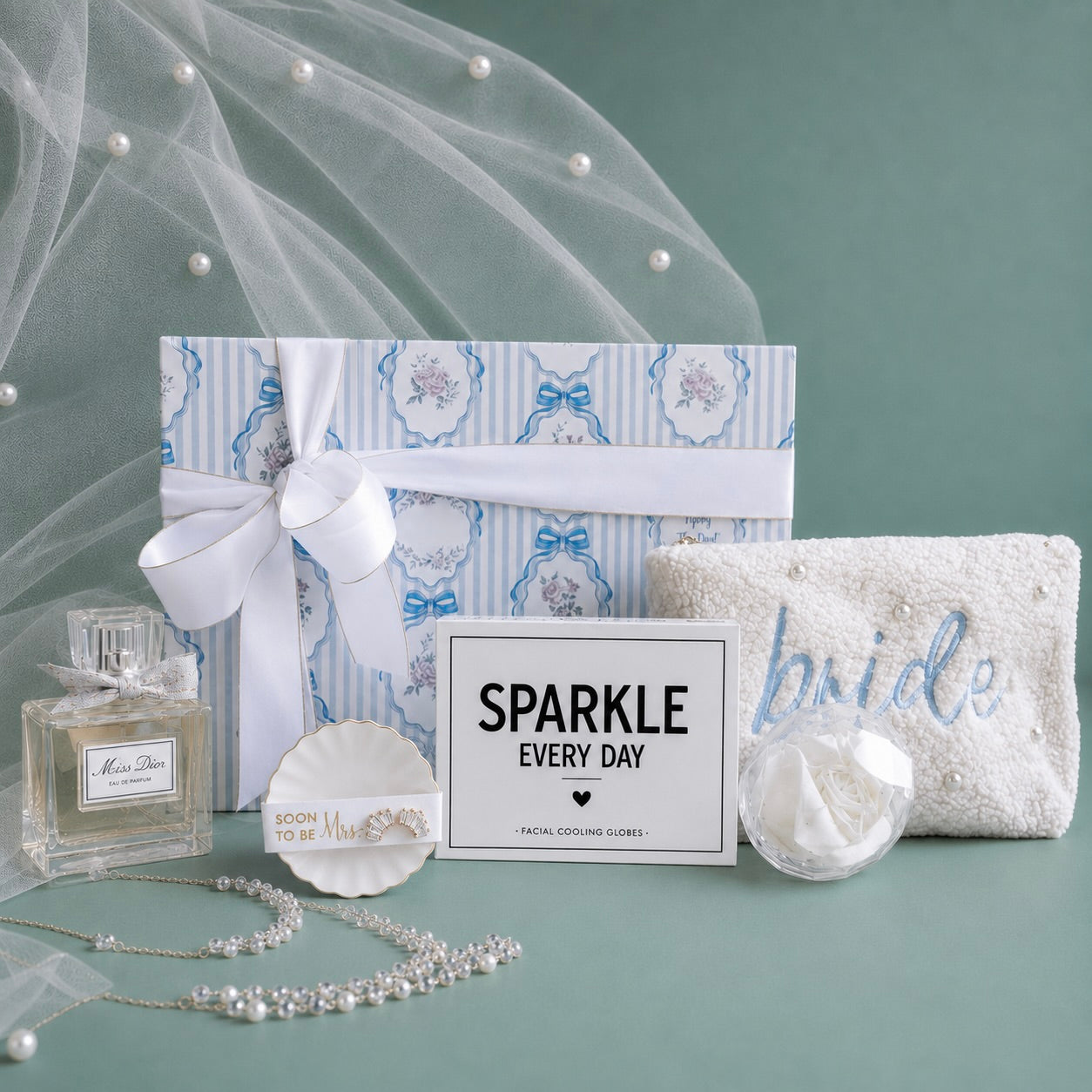 Bride shower curated gift set arranged on a sage green background, featuring a blue and white patterned gift box with a white ribbon, a white embroidered 'bride' pouch, a perfume bottle, pearl jewelry, a facial cooling globes enclosed in a 'SPARKLE EVERY DAY' box. 