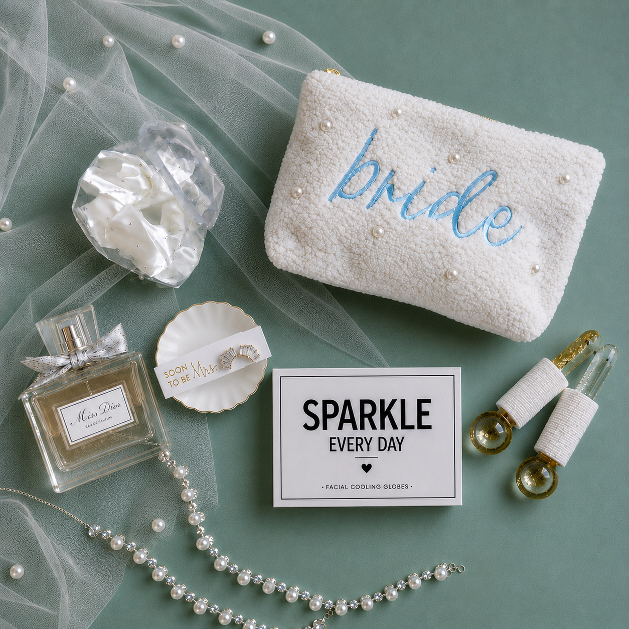 A styled flat-lay featuring a white textured pouch embroidered with the word 'bride' in blue and adorned with pearl-like beads, alongside other bridal-themed accessories including a perfume bottle, a pearl necklace, a small dish, and a facial cooling globes inclosed in 'SPARKLE EVERY DAY' box.