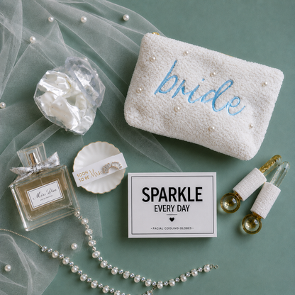 A styled flat-lay featuring a white textured pouch embroidered with the word 'bride' in blue and adorned with pearl-like beads, alongside other bridal-themed accessories including a perfume bottle, a pearl necklace, a small dish, and a facial cooling globes inclosed in 'SPARKLE EVERY DAY' box.