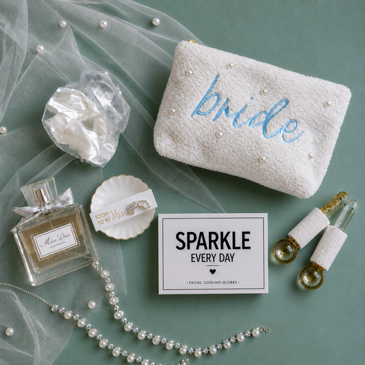 A styled flat-lay featuring a white textured pouch embroidered with the word 'bride' in blue and adorned with pearl-like beads, alongside other bridal-themed accessories including a perfume bottle, a pearl necklace, a small dish, and a facial cooling globes inclosed in 'SPARKLE EVERY DAY' box.
