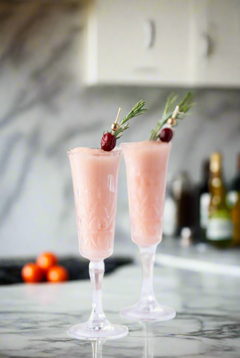 Two poinsettia cocktails in champagne flutes with garnishes on a marble countertop.