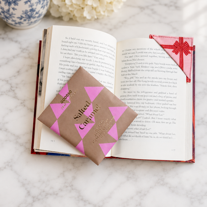 A salted caramel chocolate bar displayed on an open book. The set includes a brown paper wrap with pink geometric patterns and the words 'Salted Caramel'.