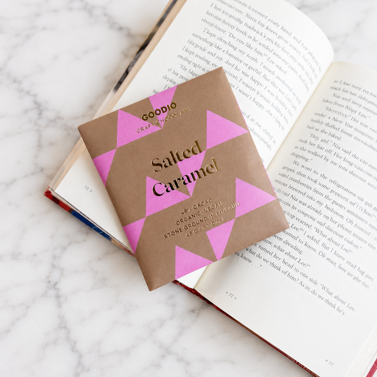 A salted caramel chocolate bar with a brown and pink geometric triangles plus gold foiling paper packaging, resting on an open book over a white marble surface.