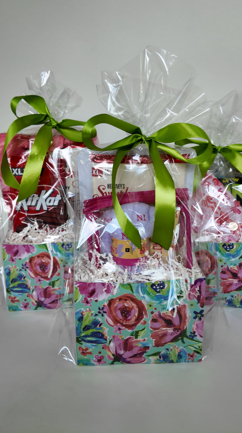 Three gift box baskets wrapped in clear cellophane with green ribbons, filled with self-care or stationery gift pieces along with assorted snacks and wrapped in floral pink and blue designed box basket.