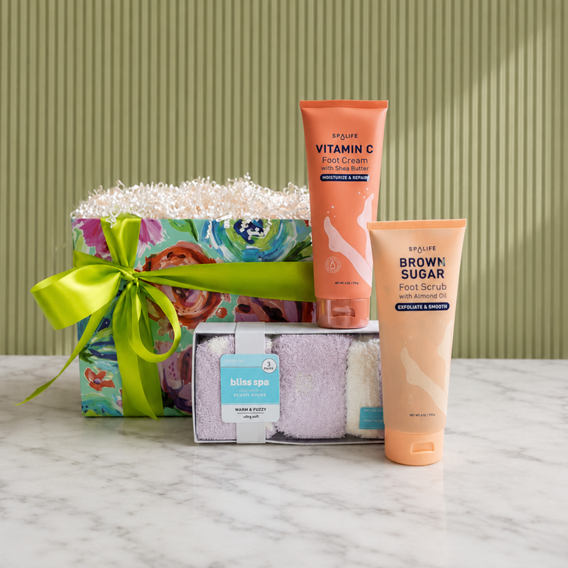 A foot spa curated gift arranged on a white marble surface against a green ribbed wall. The set includes a wrapped gift box with a bright green ribbon, three pairs of cozy socks, a vitamin V foot cream and a brown sugar with almond oil foot scrub.